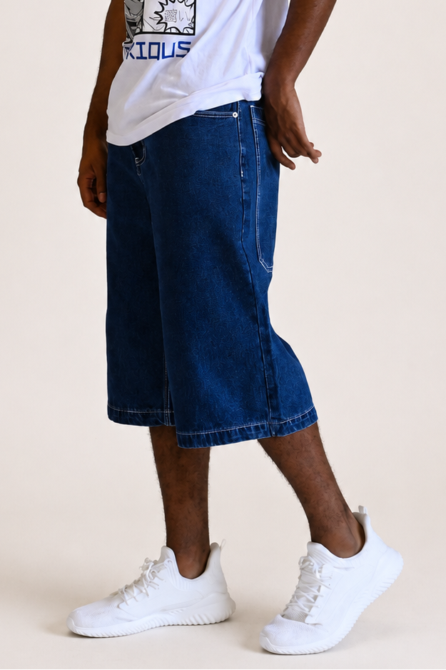 A man modeling a white graphic t-shirt with blue text and comic-style panels, paired with dark blue denim shorts and white sneakers, against a plain beige studio background.